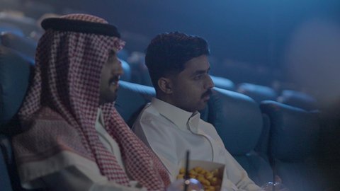 الاستمتاع بتناول الفشار أثناء مشاهدة الفلم، تصوير مقرب لصديقان عربيان سعوديان خليجيان يستمتعان بوقت الفراغ , عطلة نهاية الأسبوع , مشاهدة الأفلام والعروض المسرحية في السينما  , إيماءات وجه ويدين مختلفة