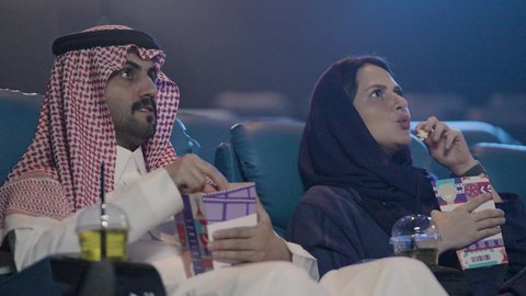 A bucket of popcorn for entertainment, a Saudi Gulf couple dressed in traditional attire watching a movie in a cinema hall, facial and hand expressions indicating excitement and enjoyment, recreational activity, screening rooms, gestures of amazement and engagement, enjoying drinks.