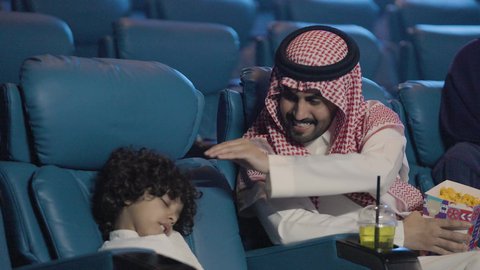 رجل  سعودي يرتدي الزي التقليدي يجلس داخل السينما برفقة ابنه يمسك بيده الفشار ، مفهوم الكسل والخمول،الشعور بالحاجة الى الراحة, تصوير مقرب لصبي سعودي نائم داخل قاعه السينما، قضاء عطله نهايه الاسبوع