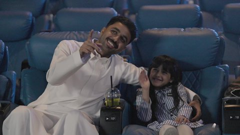 تعابير وجه و يدين تدل على الحماس و الاستمتاع ، نشاط عائلي ترفيهي، رجل خليجي يرتدي الثوب الابيض يجلس برفقه ابنته داخل القاعه ، الاستمتاع بتناول المشروبات ، ايماءات السعادة و السرور ، قضاء عطله نهايه الاسبوع