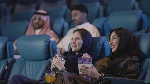 اصدقاء عربيون خليجيون سعوديون  يشاهدون فلم في احدى صالات السينما ، قضاء وقت ترفيهي سويا ، الاستمتاع بأكل دلو الفشار ، تعابير وجه و يدين تدل على الحماس و الاستمتاع ، نشاط ترفيهي ، دور العرض، الاستمتاع بتناول المشروبات