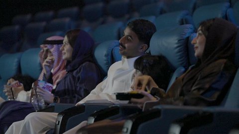 عائله عربيه خليجيه سعودية يرتدون الزي التقليدي يجلسون في صالة السينما، مشاهدة الأفلام السينمائية، عطلة نهاية الأسبوع ، قضاء أوقات ممتعة ، ايماءات السعادة و السرور ، تناول الفشار أثناء مشاهدة العروض، مشاركة الفشار، تناول المشروبات المفضلة