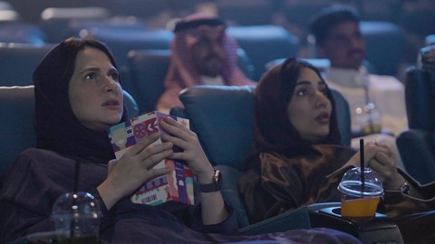 عطلة نهاية الأسبوع ، قضاء أوقات ممتعة ، مشاهدة الأفلام السينمائية ، ايماءات الاندماج و التركيز و التفاعل ، تناول الفشار أثناء مشاهدة العروض، تصوير مقرب لصديقتان عربيتان خليجيتان يرتديان الحجاب يجلسان في صالة السينما، مشاركة الفشار، تناول المشروبات المفضلة