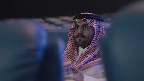 تعابير وجه و يدين تدل على الحماس و الاستمتاع ، نشاط ترفيهي ، دور العرض، متابعة الأفلام والمسرحيات على شاشة السينما، تصوير مقرب لرجل عربي خليجي سعودي يرتدي الزي التقليدي سجلس  في صالة السينما، الاستمتاع بأكل دلو الفشار