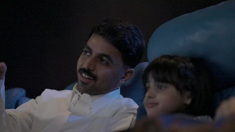 Spending enjoyable times, a close-up shot of a Saudi Arabian Gulf man wearing traditional attire sitting in a cinema hall with his son, watching the movie attentively, watching films, plays, and various shows, large screens to enhance the enjoyment of the experience, modern theaters in the Kingdom of Saudi Arabia.