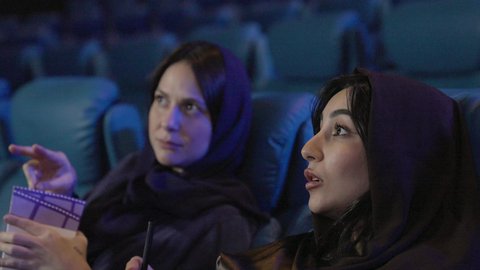 ايماءات الاندماج و التركيز، الإستمتاع بتناول الفشار، تصوير مقرب صديقتان عربيتان خليجيتان يرتديان الحجاب يجلسان في صالة السينما، قضاء أوقات ممتعة، مشاهدة الأفلام والعروض المسرحية في السينما، مشاركة الفشار، الفشار وأفلام السينما، أنشطه ترفيهيه