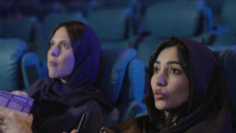 الإستمتاع بتناول الفشار، تصوير مقرب صديقتان عربيتان خليجيتان يرتديان الحجاب يجلسان في صالة السينما، قضاء أوقات ممتعة، مشاهدة الأفلام والعروض المسرحية في السينما، مشاركة الفشار، الفشار وأفلام السينما، أنشطه ترفيهيه، ايماءات الاندماج و التركيز