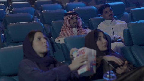 اصدقاء عربيون خليجيون سعوديون يرتدون الزي التقليدي يجلسون في صالة السينما، الإستمتاع بتناول الفشار، قضاء أوقات ممتعة، مشاهدة الأفلام والعروض المسرحية في السينما، مشاركة الفشار، الفشار وأفلام السينما، أنشطه ترفيهيه