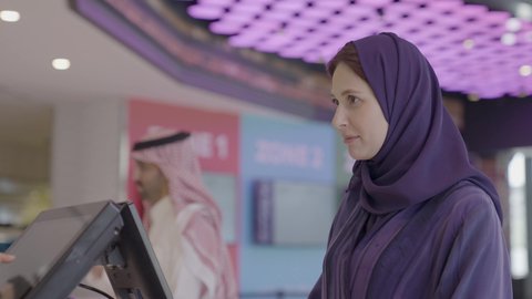 التحضير للدخول إلى قاعة السينما، سينما ومسرح، تصوير مقرب لإمرأه عربيه سعوديه خليجيه تبيع المأكولات والمشروبات في ردهة الإستقبال داخل السينما تقوم بالنقر على الشاشة، زبونه خليجيه ترتدي العباءة و الحجاب تقوم بطلب سناكس، مشاهدة الأفلام والعروض المسرحية، أنشطه ترفيهيه