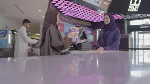Preparing to enter the screening room, a close-up shot of a Saudi Arabian Gulf woman selling food and drinks in the reception area inside the cinema, handing popcorn to customers, watching movies and theatrical performances, cinema and theater, entertainment activities.