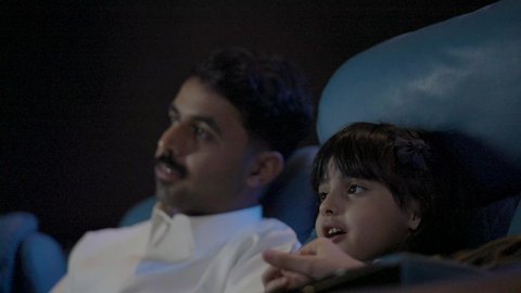 Engaging in fun recreational activities, a close-up shot of a Saudi Arabian Gulf man wearing traditional attire sitting in a cinema hall with his son, watching the movie attentively, watching films, plays, and various shows, large screens to enhance the enjoyment of the experience, modern theaters in the Kingdom of Saudi Arabia.
