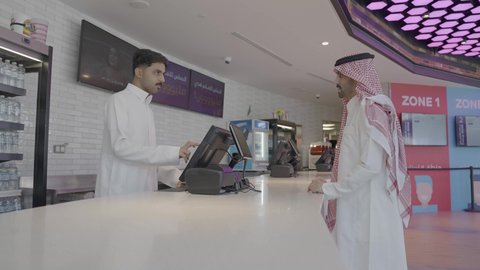 الاستعداد لبدء العرض، كاشير عربى خليجي سعودي يرتدي الثوب التقليدي يقوم بمساعدة الزبون ، رجل سعودي يرتدي الزي التقليدي يقوم بشراء سناكس المأكولات والمشروبات في ردهة الإستقبال داخل السينما، مشاهدة الأفلام والعروض المسرحية، سينما ومسرح، أنشطه ترفيهيه