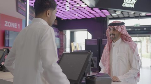Getting ready to watch the movie, a close-up shot of a Gulf Arab Saudi cashier wearing traditional attire helping a customer, a Saudi man in traditional dress buying snacks and drinks in the lobby inside the cinema, watching movies and theatrical performances, cinema and theater, recreational activities.