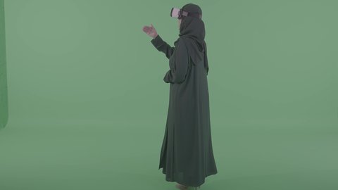 تقنيات حديثة و أجهزة تكنولوجية، قضاء وقت ممتع في اللعب، رفع الايدي عاليا بتعابير محاولة لمس شيء ما, دمج العالم الخيالي مع المتعة والتسلية، بورتريه من الخلف لامرأة عربية خليجية سعودية محجبة ترتدي نظارة الواقع الإفتراضي تقوم باللعب في ألعاب الفيديو الترفيهية, خلفية خضراء ، كروما