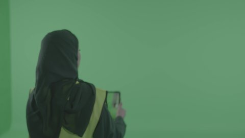 مراجعة المخططات عبر الجهاز اللوحي، مفهوم الهندسة والإنشاء المعماري, التقاط صور، تصوير مقرب من الخلف بورتريه لمهندسة عربية خليجية سعودية محجبة ترتدي العباءة والسترة تمسك بيدها التابليت، خلفية خضراء ، كروما