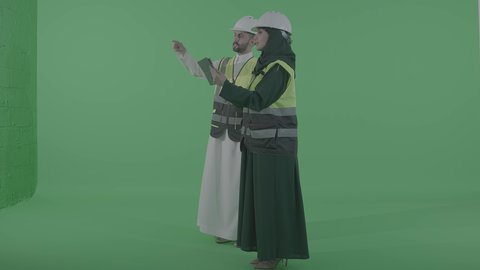 تطبيق التقنية الحديثة في إدارة المهام، مخططات وتصماميم هندسية إلكترونية، مفهوم الهندسة و الإنشاءات المعمارية, بورتريه لمهندسة عربية خليجية سعودية ترتدي الخوذة وسترة الحماية تحمل بيدها جهاز التابلت تتحدث مع رجل عربي خليجي سعودي، الجسم بالطول الكامل، خلفية خضراء ، كروما