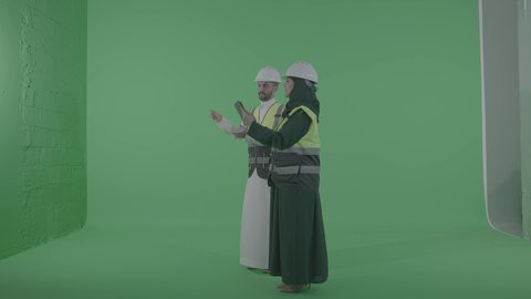 دمج العمل مع التقنية و التكنولوجيا، مفهوم الهندسة و الإنشاءات المعمارية, مخططات وتصماميم هندسية إلكترونية, بورتريه لمهندسة عربية خليجية سعودية ترتدي الخوذة وسترة الحماية تحمل بيدها جهاز التابلت تتحدث مع رجل عربي خليجي سعودي، الجسم بالطول الكامل، خلفية خضراء ، كروما