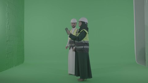 مفهوم الهندسة و الإنشاءات المعماريه, دمج العمل مع التقنية و التكنولوجيا, مخططات وتصماميم هندسية إلكترونية, بورتريه لمهندسة عربية خليجية سعودية ترتدي الخوذة وسترة الحماية تحمل بيدها جهاز التابلت تتحدث مع رجل عربي خليجي سعودي، الجسم بالطول الكامل، خلفية خضراء ، كروما