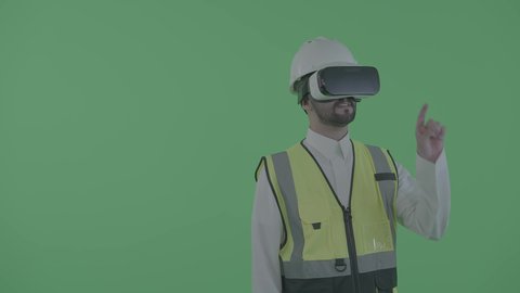 Interaction with digital engineering plans, the growth of the technological engineering field, working in the engineering sector through virtual reality technology, a portrait of a Saudi Gulf Arab engineer wearing a work vest, helmet, and VR glasses interacting with a holographic building plan model, green background, chroma.