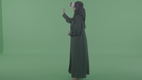 التفاعل مع العناصر الافتراضية باللمس، بورترية من الجانب لامراة عربيه خليجيه سعودية محجبه ترتدي العباءة والنظارة نظارات ثلاثية الأبعاد (الفي ار) ، تعابير النقر على شاشة ، محاكاة العالم الخيالي، خلفية خضراء ، كروما