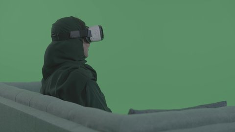 Enjoying leisure time, using modern electronic technologies and devices, a portrait of a young Arab Gulf Saudi woman wearing a hijab sitting on a couch wearing 3D VR glasses, immersing and interacting with the virtual world, green background, chroma.