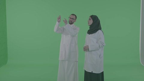 Saudi work environment, a portrait of an Arab Gulf doctor and doctor wearing white medical coats discussing medical matters, clicking and pointing at something, green background, chroma.