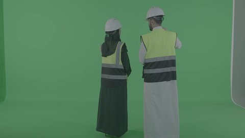 الإشراف والتدقيق على العمل، مهن ووظائف سعودية، بورترية من الخلف لمهندس عربيى خليجي سعودي يقوم بالتعاون ومتابعة الاعمال والمخططات برفقة زميلتة، ارتداء سترة وخوذة الامان والحماية، خلفية خضراء ، كروما