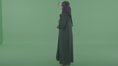 Spending enjoyable time playing, raising hands high with expressions of trying to touch something, modern technologies and devices, merging the imaginary world with fun and entertainment, a portrait from behind of a Saudi Arabian Gulf woman wearing a hijab and virtual reality glasses playing video games, green background, chroma.