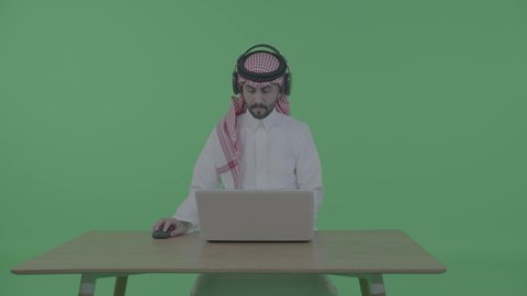 تصفح الإنترنت والعمل عن بعد، إنجاز مهام العمل من خلال اللابتوب، بورتريه لرجل عربي خليجي سعودي يرتدي الثوب التقليدي والشماغ و سماعات الرأس يجلس على الطاولة و يحمل بيده جهاز الحاسوب المحمول، الضغط على أزرار لوحة المفاتيح، اجراء مكالمه عمل فيديو ، خلفية خضراء ، كروما