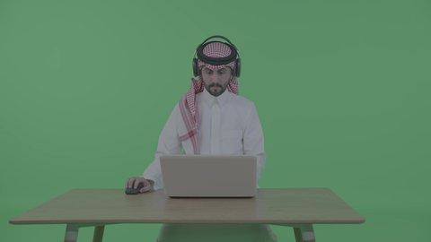 إنجاز مهام العمل من خلال اللابتوب، بورتريه لرجل عربي خليجي سعودي يرتدي الثوب التقليدي والشماغ و سماعات الرأس يجلس على الطاولة و يحمل بيده جهاز الحاسوب المحمول، تصفح الإنترنت والعمل عن بعد، الضغط على أزرار لوحة المفاتيح، اجراء مكالمه عمل فيديو ، خلفية خضراء ، كروما