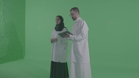 تقديم الاستشارات للمرضى عن بعد الأجهزة التكنولوجية، دمج التقنيات الحديثة بالمجالات الطبية, تطور القطاع الصحي في المملكة العربية السعودية، بورتريه مقرب لطبيب عربي خليجي سعودي يرتدي المعطف الأبيض يمسك جهاز التابلت بيديه برفقة مساعدته، خلفية خضراء ، كروما