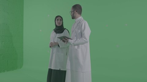 دمج التقنيات الحديثة بالمجالات الطبية, تطور القطاع الصحي في المملكة العربية السعودية، تقديم الاستشارات للمرضى عن بعد الأجهزة التكنولوجية، بورتريه مقرب لطبيب عربي خليجي سعودي يرتدي المعطف الأبيض يمسك جهاز التابلت بيديه برفقة مساعدته، خلفية خضراء ، كروما