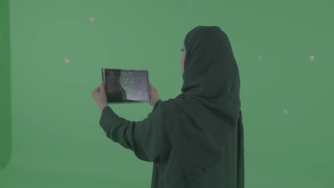 إجراء مكالمات فيديو عبر الإنترنت، بورترية من الخلف لإمرأه عربيه سعوديه خليجيه محجبة ترتدي العباءة تمسك بيديها جهاز التابلت, انجاز مهام العمل عن بعد, مشاهدة فلم, تسليه وترفيه, قضاء وقت ممتع, خلفيه خضراء، كروما