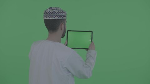متابعه أخر أخبار مواقع التواصل الاجتماعي ، بورتريه من الخلف لرجل عربي خليجي عماني يرتدي الدشداسة و الكمه يقوم بالنقر على جهاز التابلت بشاشه خضراء فارغه، التواصل والدردشه مع الأقارب والأصدقاء، التسويق الالكتروني عن بعد، خلفية خضراء، كروما
