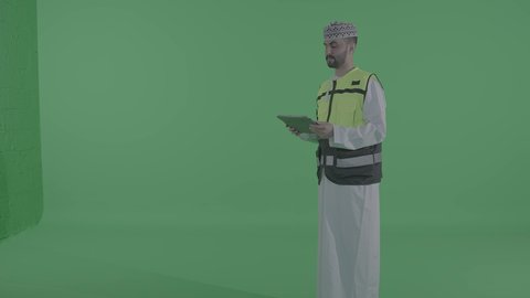 Production and manufacturing efficiency and achieving the industrial revolution, adopting modern technologies and technology in engineering, an Arab Gulf Omani engineer wearing a dishdasha, kumma, jacket, and safety helmet monitoring the factory operations using a tablet, Omani professions and jobs, green background, chroma.