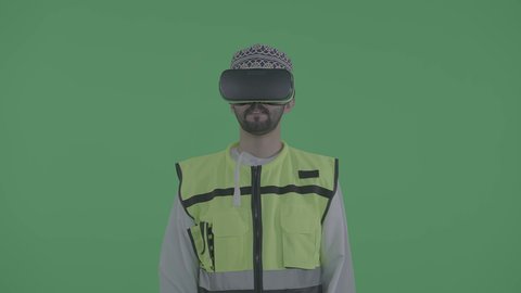 تجسيد ومشاهدة المشاريع الهندسية بالاعتماد على التكنولوجيا، الاندماج مع الهندسة الافتراضية، بورتريه لمهندس عربي خليجي عماني يرتدي الدشداشة و الكمة والسترة الخاصة بالعمل ونظارة الواقع الافتراضي يرفع يديه للمس عناصر هولوجرامية، خلفية خضراء ، كروما