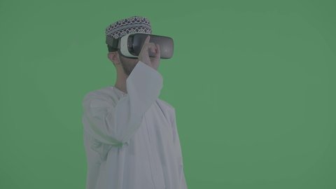 المتعة والتسلية بالمشاهدة عبر نظارة الواقع الافتراضي، التطور التقني والتكنولوجي، بورتريه لشاب عربي خليجي عماني يرتدي الدشداشة والكمة ونظارات ثلاثية الأبعاد (الفي ار) يرفع يده للمس عناصر هولوجرامية, الاندماج ومحاكاة العالم الخيالي،خلفية خضراء ، كروما