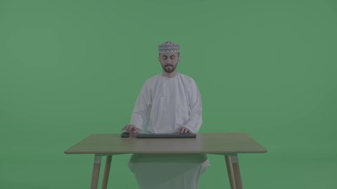 بيئة العمل في المكاتب والشركات العمانية، دمج التقنيات الحديثة بالمجال الإداري والمكتبي، تصوير لرجل عربي خليجي عماني يرتدي الدشداشة و الكمه يجلس على طاولة المكتب يقوم إدخال البيانات والكتابة من خلال لوحة المفاتيح ، خلفية خضراء ، كروما