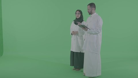 استخدام التقنيات المتطورة في المجال الطبي، تقديم الاستشارات للمرضى عن بعد، بورتريه لطبيب عربي خليجي سعودي يرتدي المعطف الابيض يحمل جهاز التابلت بيده برفقة مساعدته ،  الجسم بالطول الكامل ، خلفية خضراء، كروما