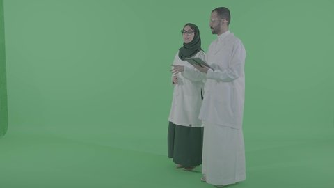 دمج التقنيات الحديثة بالمجال الطبي، تقديم الاستشارات للمرضى عن بعد، بورتريه لطبيب عربي خليجي سعودي يرتدي المعطف الابيض يحمل جهاز التابلت بيده برفقة مساعدته ،  الجسم بالطول الكامل ، خلفية خضراء، كروما