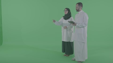 الجمع بين الرعاية الطبية والتكنولوجيا الحديثة، بورتريه لطبيب عربي خليجي سعودي يرتدي المعطف الابيض يحمل جهاز التابلت بيده برفقة مساعدته ، طبيبة سعودية تقوم بالرسم والتخطيط في الهواء، تعابير النقر على شاشة ، استخدام حاسة اللمس لتحريك العناصر الهولوجرامية، خلفية خضراء، كروما
