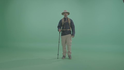 Looking at the camera with gestures of happiness and joy, a portrait of an Arab Gulf Saudi explorer wearing suitable mountain clothing for mobility and travel, holding a walking stick, ready for tourist adventures, outdoor adventure and camping activities, full-body shot, green background, chroma.