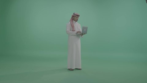 انجاز وادارة المهام عبر الأونلاين، النقر على لوحة المفاتيح ، مفهوم العمل عن بعد, دمج العمل مع التقنية والتكنولوجيا الحديثة والمتطورة, بورتريه لشاب عربي خليجي سعودي يرتدي الثوب التقليدي والشماخ يستخدم الحاسب المحمول, التواصل والدردشة مع الأصدقاء والأهل عبر السوشال ميديا، الجسم بالطول الكامل، خلفية خضراء، كروما