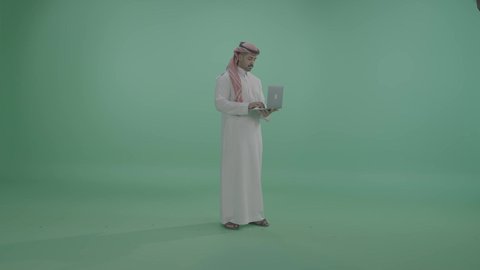 النقر على لوحة المفاتيح ،مفهوم العمل عن بعد, دمج العمل مع التقنية والتكنولوجيا الحديثة والمتطورة, انجاز وادارة المهام عبر الأونلاين, بورتريه لشاب عربي خليجي سعودي يرتدي الثوب التقليدي والشماخ يستخدم الحاسب المحمول, التواصل والدردشة مع الأصدقاء والأهل عبر السوشال ميديا، الجسم بالطول الكامل، خلفية خضراء، كروما