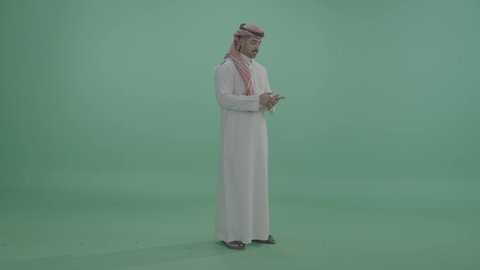 التواصل مع الاهل و الاصدقاء, ايماءات السعادة و السرور، بورتريه لشاب عربي خليجي سعودي يرتدي الزي التقليدي و الشماغ  يقوم بإجراء مكالمة هاتفية عبر الهاتف المحمول، استخدام التقنيات الحديثة المتطورة, صورة الجسم بالطول الكامل, خلفية خضراء ، كروما