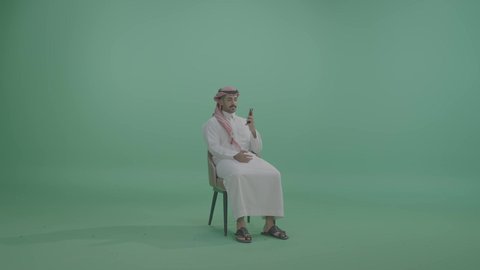 The use of advanced modern technologies, communication with family and friends, gestures of happiness and joy, a portrait of a young Arab Gulf Saudi man wearing traditional attire and a ghutrah sitting on a chair making a phone call via a mobile phone, green background, chroma.
