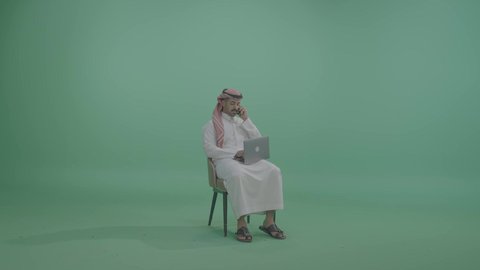 اجراء مكالمة هاتفية عبر الهاتف المحمول، مفهوم العمل عن بعد, دمج العمل مع التقنية والتكنولوجيا الحديثة والمتطورة, انجاز وادارة المهام عبر الأونلاين, بورتريه لشاب عربي خليجي سعودي يرتدي الثوب التقليدي والشماخ يجلس على كرسي يستخدم الحاسب المحمول، خلفية خضراء، كروما