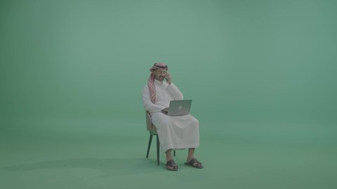 التواصل والدردشة مع الأصدقاء والأهل عبر الهاتف المحمول، مفهوم العمل عن بعد, دمج العمل مع التقنية والتكنولوجيا الحديثة والمتطورة, انجاز وادارة المهام عبر الأونلاين, بورتريه لشاب عربي خليجي سعودي يرتدي الثوب التقليدي والشماخ يجلس على كرسي يستخدم الحاسب المحمول، خلفية خضراء، كروما