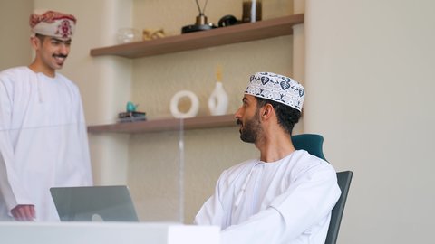 الحوارات والنقاشات ضمن الفريق، رجل عربي خليجي عماني يرتدي الدشداشة و الكمه يجلس فى مكتبة يستخدم الحاسب المحمول و يقوم بمصافحة زميله،  السلام والتحية والاتفاق على انجاز المهام