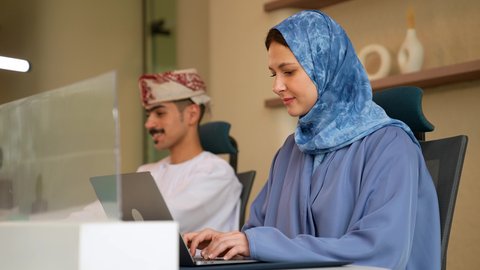 مفهوم الشراكة والتعاون, بيئة العمل العماني ، امراة عربيه خليجيه عمانية محجبه ترتدي العباءة تجلس داخل مقر الشركة برفقة زميلها تقوم باستخدام الحاسب المحمول و الكتابة على لوحه المفاتيح ، ايماءات الاندماج و التركيز